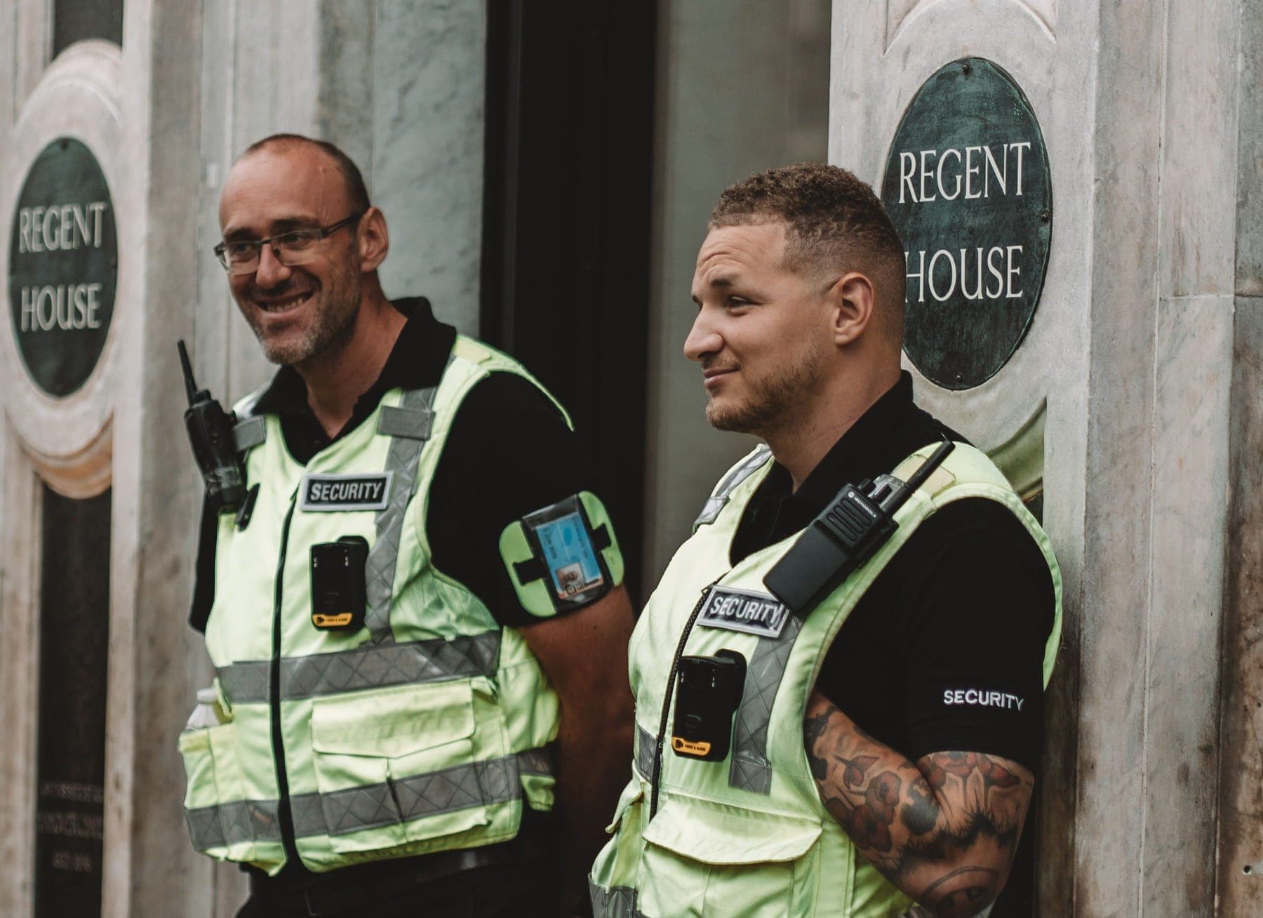 Two smiling SIA-licensed security officers in hi-vis vests with radios at a building entrance